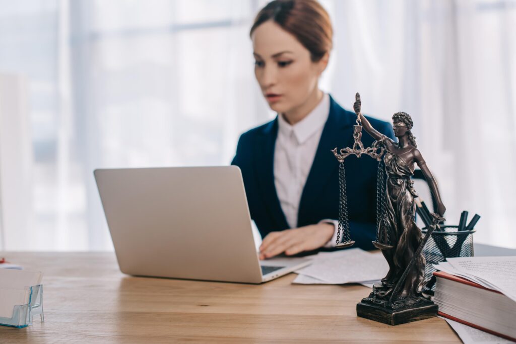 selective focus of female lawyer in suit working on laptop at workplace with femida in office